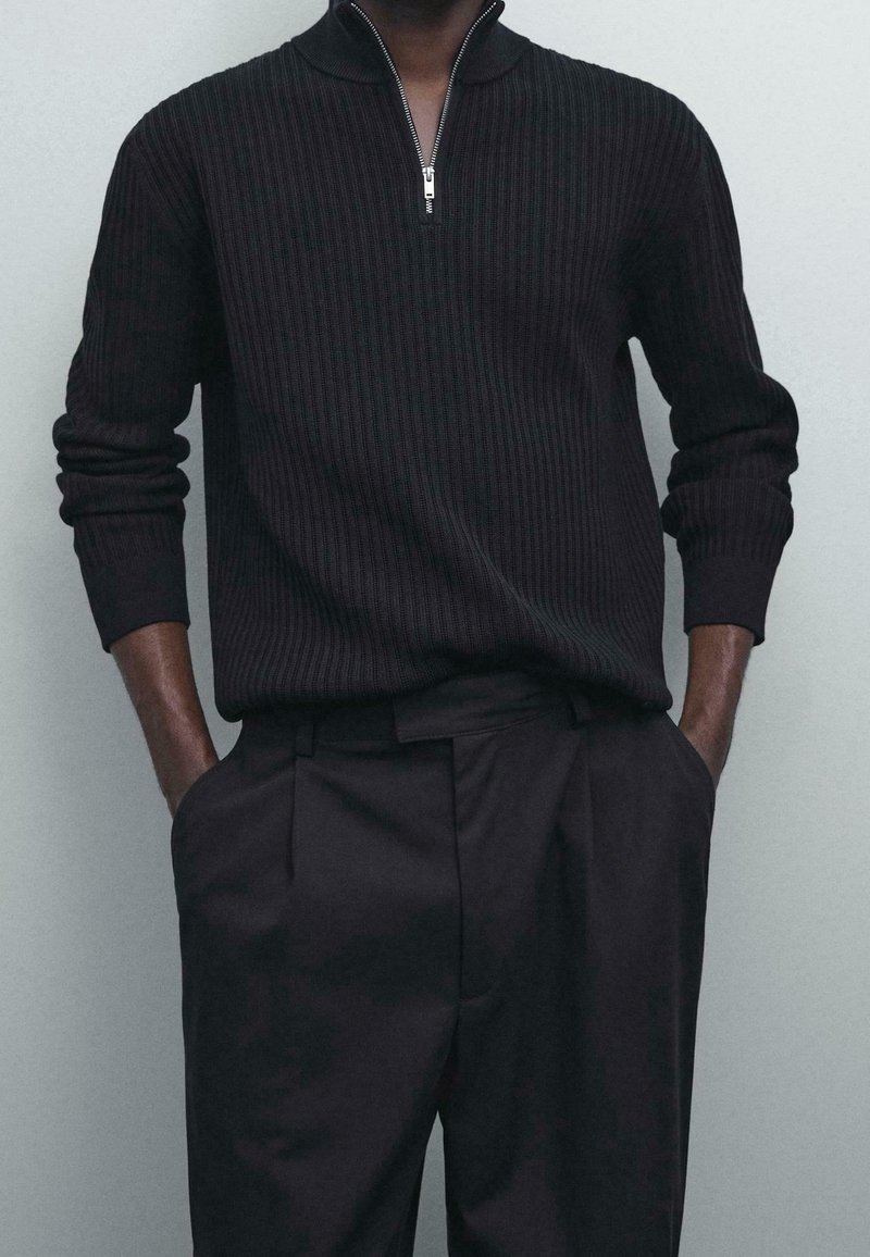 Homme portant un pull côtelé noir avec une demi-fermeture éclair et un pantalon noir, mains dans les poches, sur un fond clair uni.