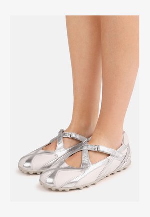 Voeten die zilver-witte gesloten Mary Jane-schoenen met gekruiste bandjes en rubberen zolen dragen op een lichte achtergrond.