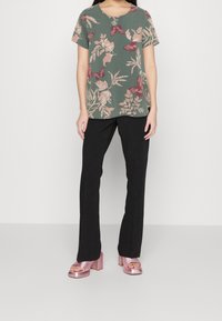 Mujer vestida con blusa verde con estampado de mariposas y flores, pantalones negros de pierna recta y sandalias de tacón bloque metálico rosa, de pie contra un fondo blanco.