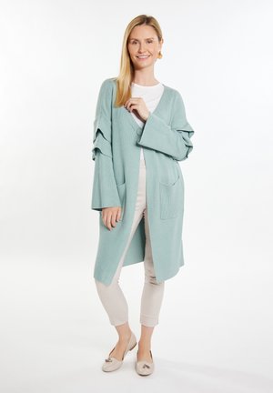 Cardigan lung de culoare verde deschis, cu decolteu în V, manșete răsucite și buzunare frontale. Purtați peste un top alb și asortat cu pantaloni bej și pantofi.