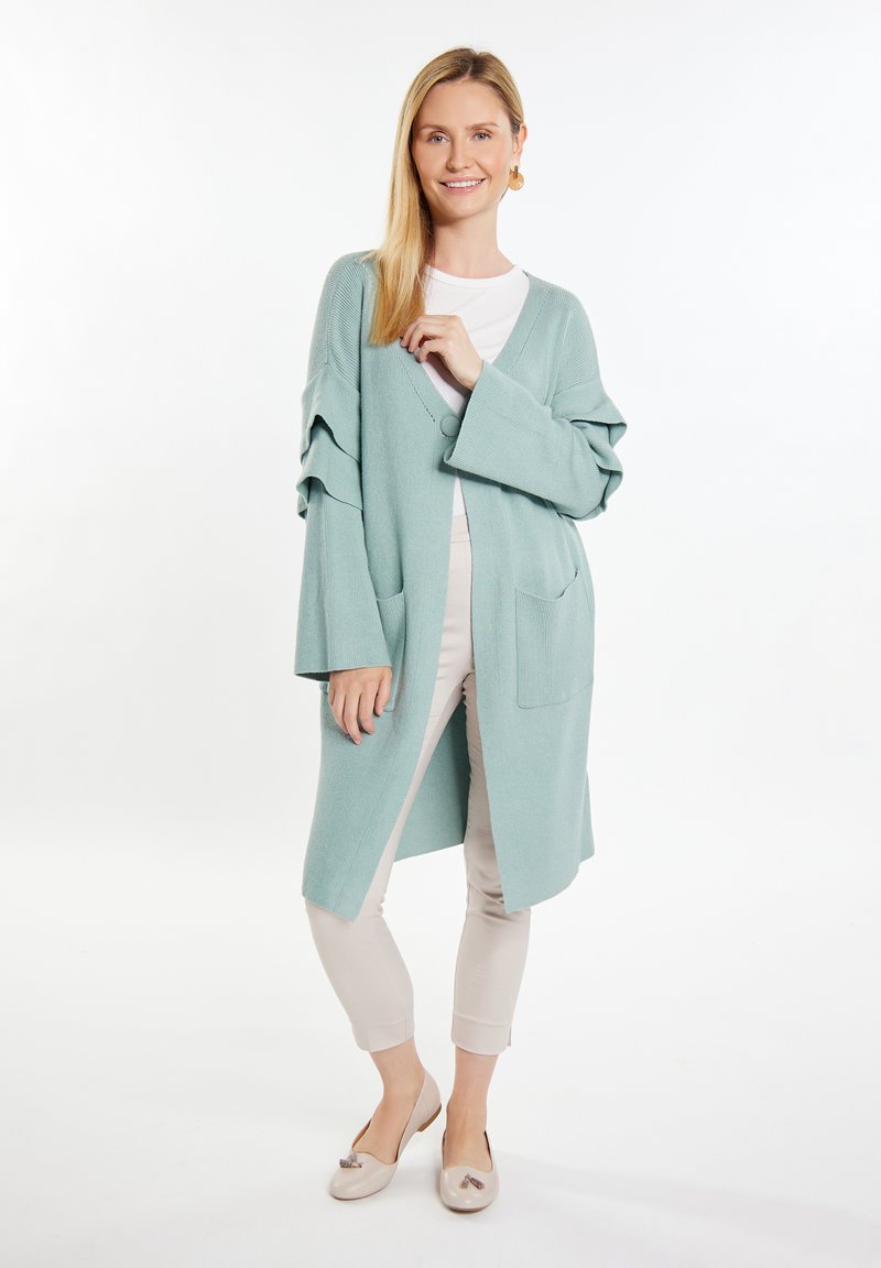 Cardigan lungo verde chiaro con scollo a V, maniche arrotolate e tasche frontali. Indossato sopra una maglietta bianca e abbinato a pantaloni e scarpe beige.