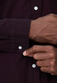 Dunkellilahem Hemd mit weißen Knöpfen, mit einem spitzen Kragen und einem strukturierten Stoff. Eine Hand ist dabei, die Manschette zuzuknöpfen.