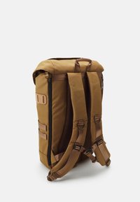 Sac à dos en tissu marron avec un design à rabat enroulable, bretelles rembourrées, fermeture éclair latérale et sangles externes pour attacher des accessoires. Dos en maille pour plus de confort.