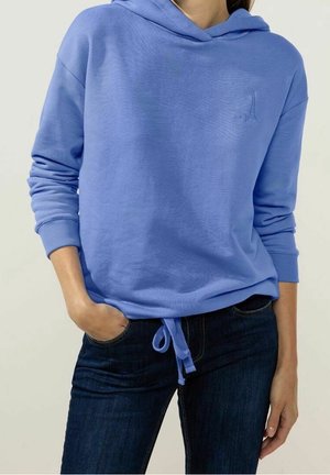 Woman wearing a light blue hooded sweatshirt with an Eiffel Tower embroidery and dark blue jeans, with one hand in pocket.