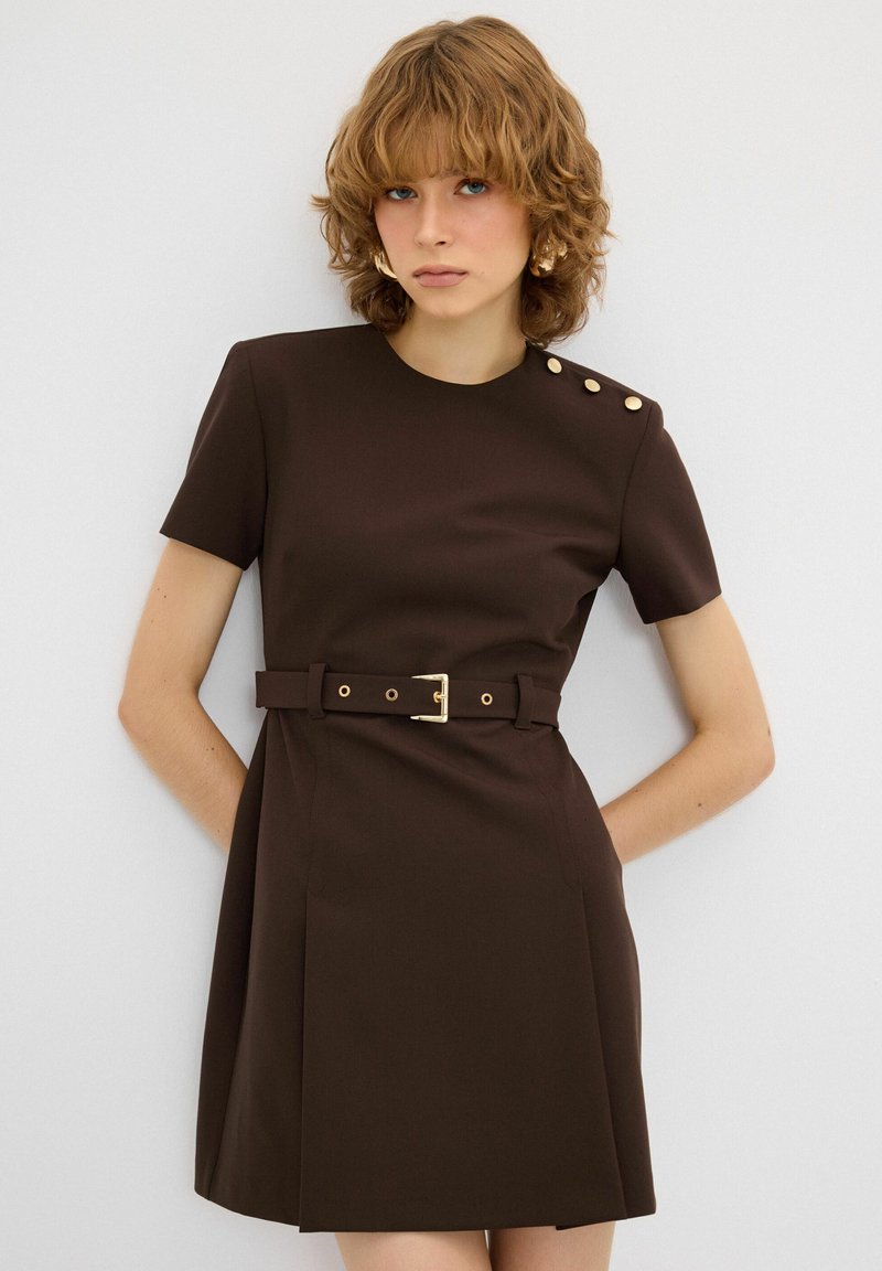 Bruine jurk met korte mouwen, een taille met riem, zijplooien en gouden knoopaccenten op de schouder. Glad stoftextuur.
