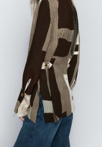 Chemise à manches longues présentant un design patchwork aux tons marron, crème et vert, avec des motifs rayés et un détail de bouton sur le poignet.