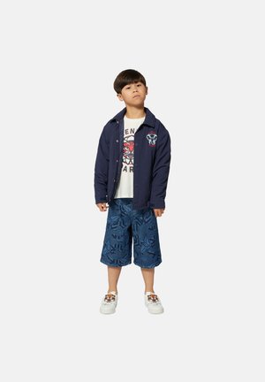 Marineblauwe jas met logo, wit t-shirt met grafische druk, blauwe gebloemde shorts en witte schoenen met tijgerontwerp.