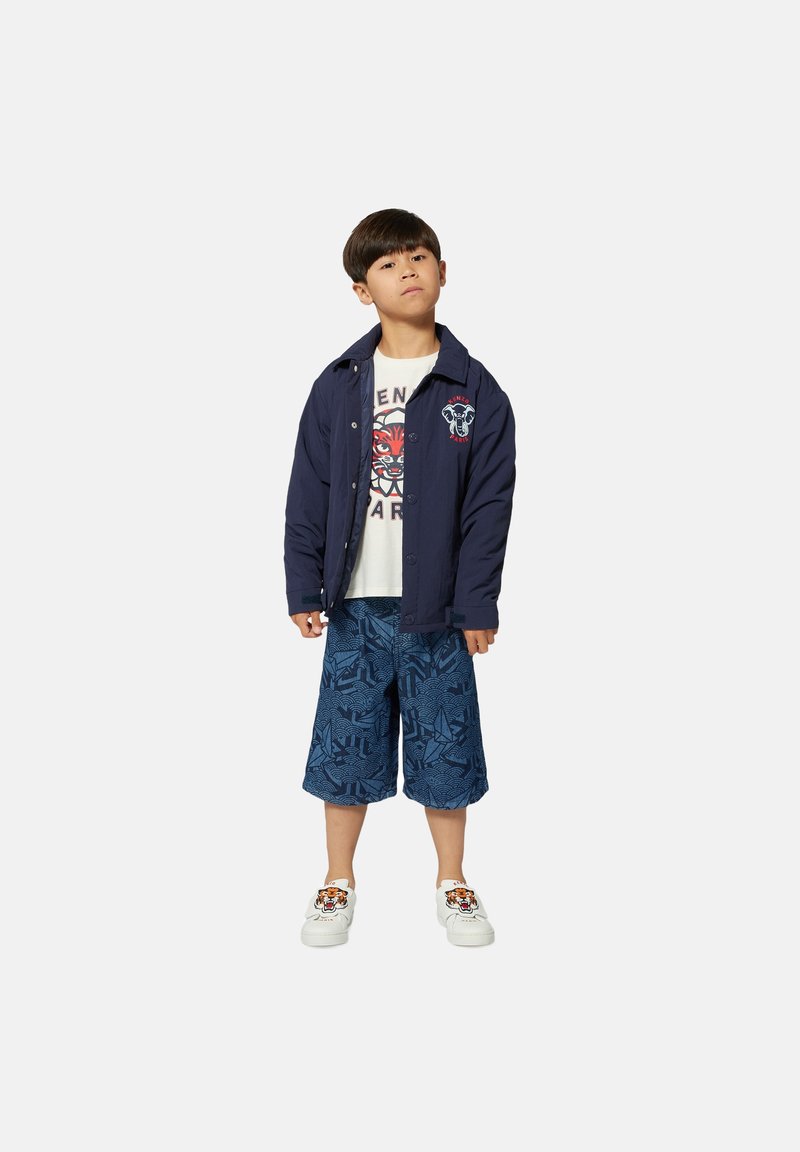 Marineblauwe jas met logo, wit t-shirt met grafische druk, blauwe gebloemde shorts en witte schoenen met tijgerontwerp.