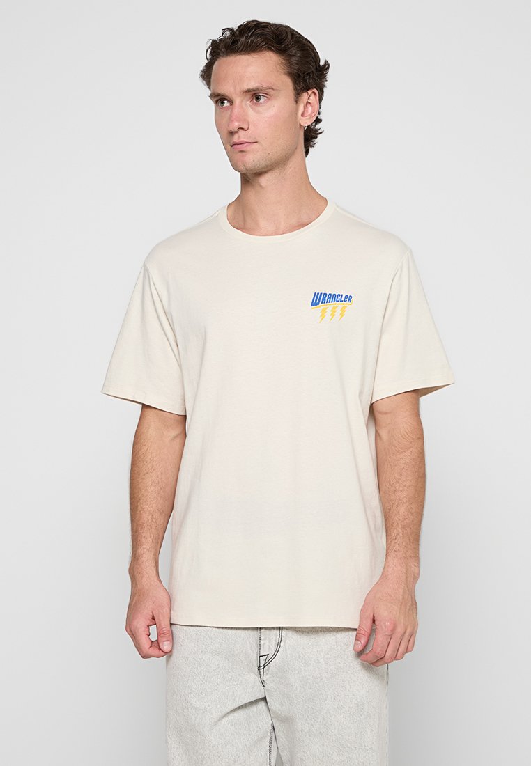 Camiseta de manga corta beige en algodón suave con cuello redondo. Presenta un logo "Wrangler" en azul y amarillo en el pecho. Ajuste casual.