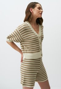 Gestreifte beige und cremefarbene Strickjacke mit V-Ausschnitt, kurzen Ärmeln und Knopfverschluss, kombiniert mit passenden Shorts im gleichen Muster.