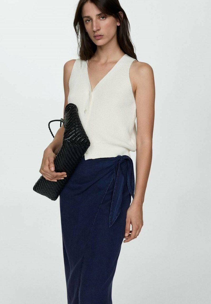 Haut en tricot sans manches beige clair avec décolleté en V, associé à une jupe portefeuille bleu marine. Tient un sac à main tissé noir texturé. Design simple et moderne.