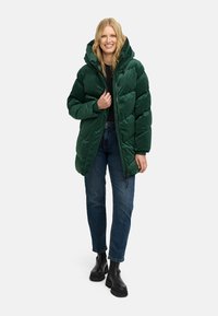 Chaqueta de plumón verde con capucha, que cuenta con un cierre de cremallera y diseño acolchado. Combinada con jeans azul oscuro y botas negras de tobillo.