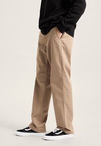 Pantaloni beige in cotone con una vestibilità morbida e tasche laterali, abbinati a sneakers bianche e nere con lacci. La camicia è nera con maniche lunghe.