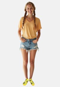Gelbes V-Ausschnitt-T-Shirt, zerrissene Denim-Shorts mit fransigen Kanten und leuchtend gelbe Socken mit grünen Avocado-Mustern.