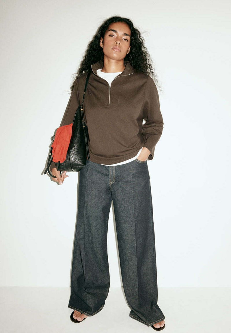 Femme aux longs cheveux bouclés portant un haut marron zippé, un jean large foncé et des sandales noires, tenant un sac noir avec un tissu orange.