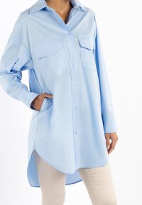 Camicia azzurra a maniche lunghe con due tasche sul petto, chiusura con bottoni e orlo curvo. Realizzata in tessuto morbido con una leggera lucentezza.