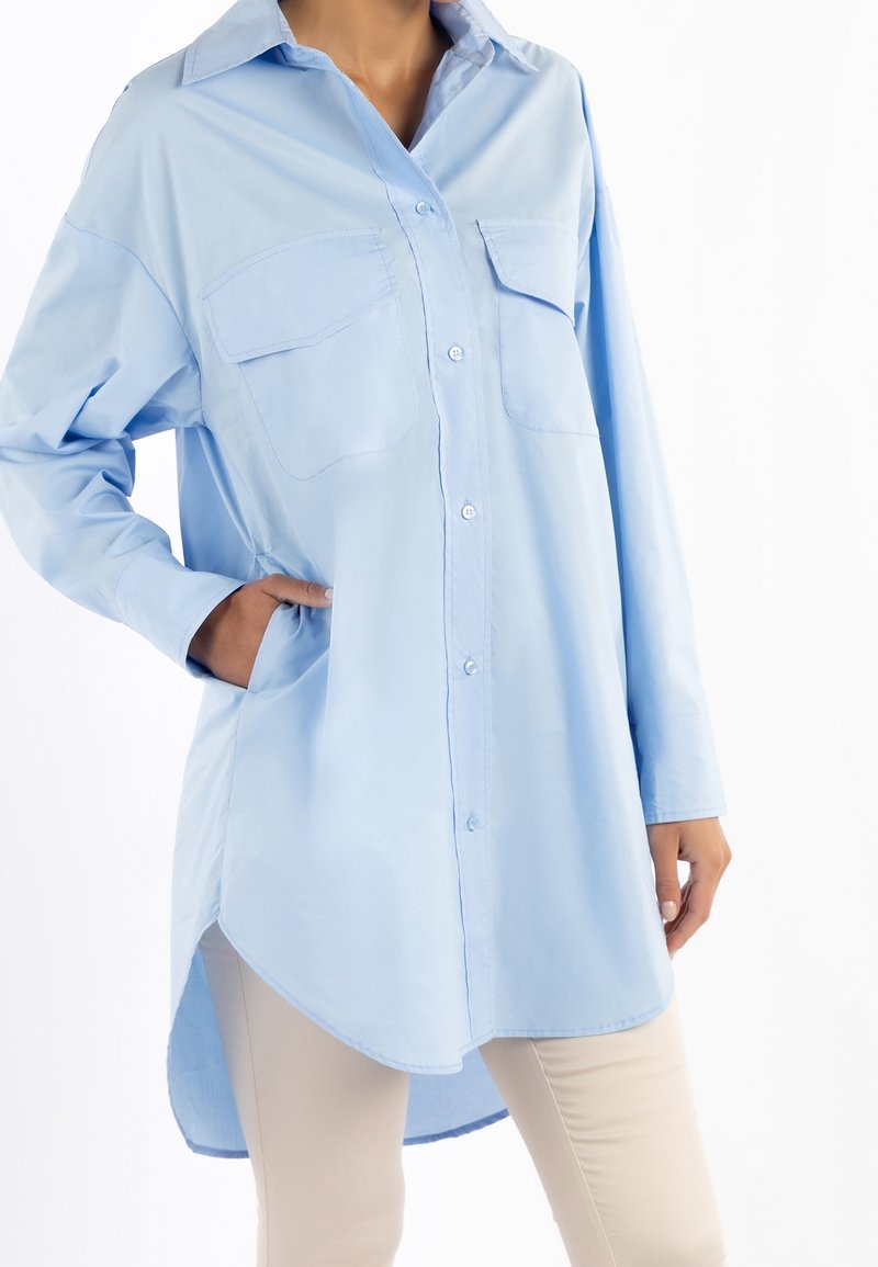 Camicia azzurra a maniche lunghe con due tasche sul petto, chiusura con bottoni e orlo curvo. Realizzata in tessuto morbido con una leggera lucentezza.
