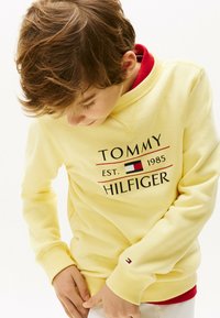 Sudadera amarilla con puños y cuello acanalados, que presenta el logo "TOMMY HILFIGER" en negro y rojo, superpuesta sobre una camiseta roja.