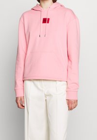 Sweat à capuche rose en tissu doux, avec une poche avant, une capuche à cordon de serrage, et un patch logo rouge sur la poitrine. Manches longues avec poignets.