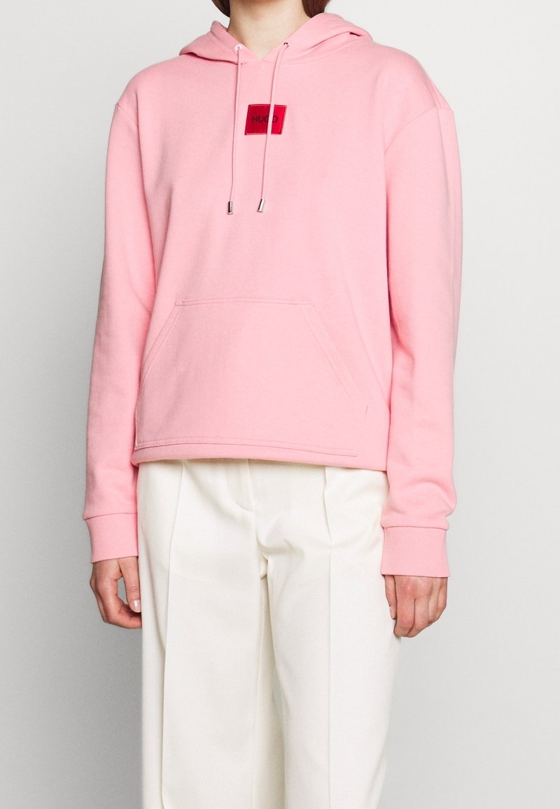 Sweat à capuche rose en tissu doux, avec une poche avant, une capuche à cordon de serrage, et un patch logo rouge sur la poitrine. Manches longues avec poignets.