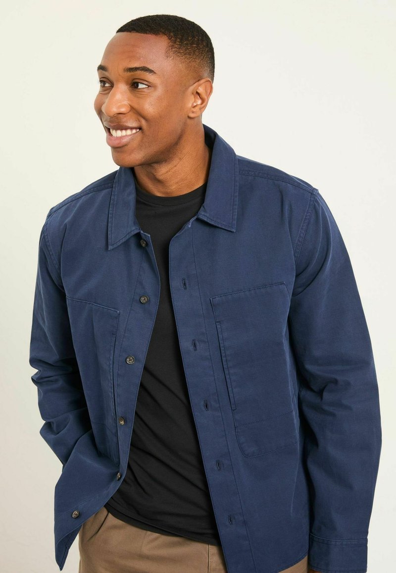 Tmavomodrá bunda na gombíky s golierom košele, predným vreckom a dlhými rukávmi, nosená cez čierne tričko. Hladká textúra.