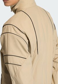 Μπεζ μπουφάν από ελαφρύ υλικό, με διακριτικές μαύρες ραφές, ψηλό γιακά και άνετη εφαρμογή με ανάγλυφες λεπτομέρειες.