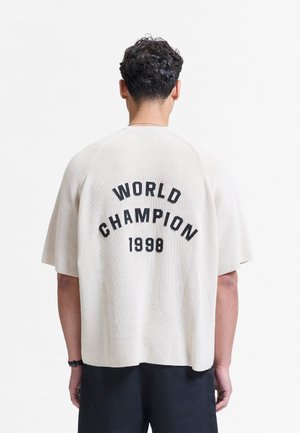 Mand med krøllet hår står med ryggen til iført en løs hvid strikket top med "WORLD CHAMPION 1998" i sorte bogstaver på ryggen.