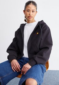 Hoodie noir zippé avec poches avant, poignets côtelés et un petit logo orange sur la poitrine. Porté par-dessus un t-shirt blanc et associé à un short en denim bleu.
