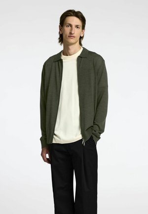 Oliven grøn zip-jakke over en cremefarvet t-shirt, parret med sorte bukser. Glat tekstur og afslappet design med krave.