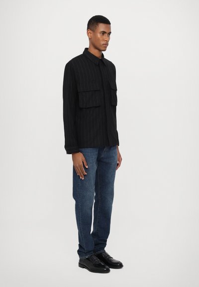 Svart skjorta med randigt mönster och två bröstfickor, matchad med blå jeans och svarta skor. Skjortan har en krage och knappstängning.
