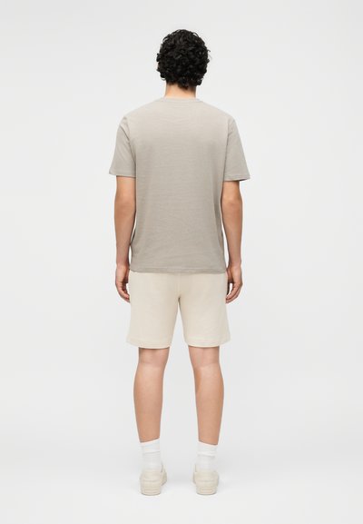 Άνδρας με σγουρά σκούρα μαλλιά, όρθιος γυρισμένος με την πλάτη, φοράει μπεζ κοντομάνικο πουκάμισο, ανοιχτόχρωμο σορτς, λευκές κάλτσες και μπεζ αθλητικά παπούτσια.