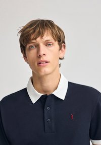 Navyblaues Poloshirt mit weißem Kragen, zwei Knöpfen und rotem Logo auf der Brust. Der Stoff wirkt glatt und atmungsaktiv.