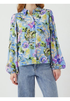 Kvinde iført blå blomstret bluse med pufærmer kombineret med lyseblå cowboybukser, stående mod en ensfarvet baggrund.