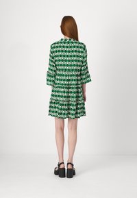 Vihreä kuviollinen mekko, jossa on kolmiomaisia motiiveja, röyhelöhihoja ja kerrostettu helma. Yhdistetty mustiin platform-sandaaleihin.