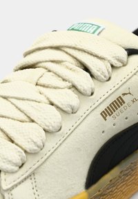 Puma suede sneakers in cream and black with textured materials. Features thick, woven cream laces and a green logo label on the tongue.