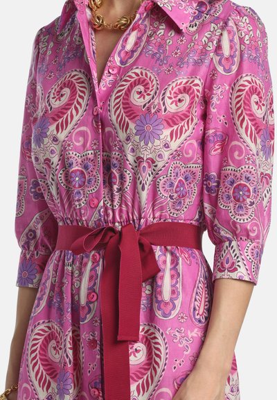 Rosa kjole med paisley- og blomsterprint, knapper foran, trekvartlange ærmer og et mørkerødt bælte bundet i taljen, båret med en guldkæde.