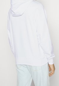 Sudadera blanca con cordón y un gran bolsillo trasero, presentada con un corte relajado y puños acanalados. Combinada con pantalones de mezclilla claros.