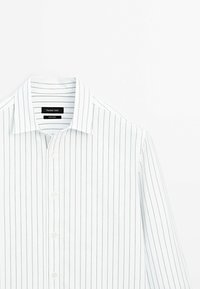 Chemise blanche à manches longues avec de fines rayures verticales noires, boutonnée à l'avant et avec un col, présentée sur un fond blanc.