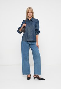 Denimskjorte med krage og knappelukking, sammen med vide blå jeans. Tilbehør inkluderer en liten mørk håndveske og svart høye hæler.