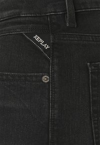 Jeans de mezclilla negra con un pequeño logo bordado "REPLAY" en el bolsillo trasero, herrajes de botón plateados y una textura suave.