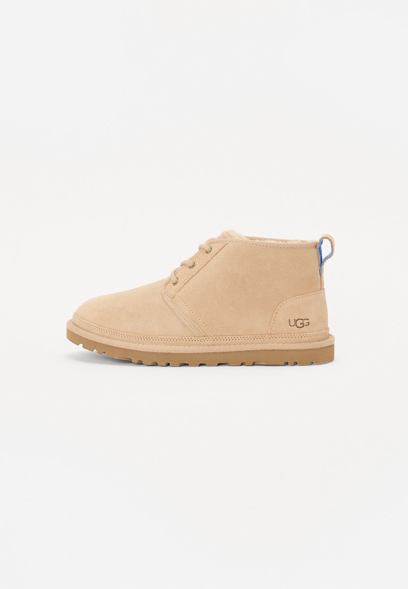 Botines de ante color beige con puntera redonda, cordones y suela de goma corta. Presentan un acento en el tacón azul y el logotipo de UGG en el lateral.