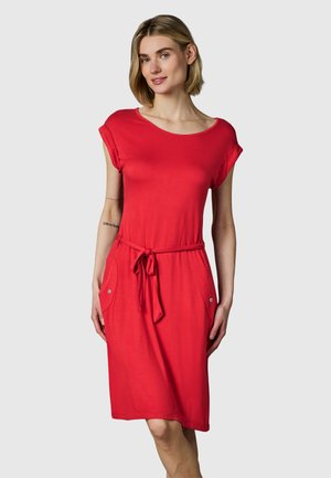 Donna che indossa un vestito rosso lungo fino al ginocchio con maniche corte arrotolate, cintura con fiocco in vita e dettagli delle tasche su sfondo grigio chiaro.