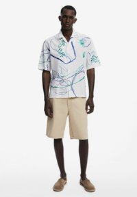 Camicia bianca a maniche corte con design astratti blu e verde, abbinata a pantaloni corti beige e scarpe slip-on marroni. Tessuto dal tocco morbido.