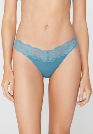 Buste féminin portant des sous-vêtements bleu sarcelle ornés de dentelle et un soutien-gorge assorti, debout avec les bras détendus le long du corps sur un fond uni.