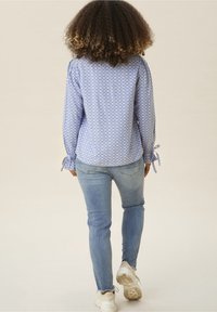 Blusa blu con motivo a fantasia, maniche lunghe, polsini annodati e tessuto leggero, abbinata a jeans blu chiaro e sneakers bianche.