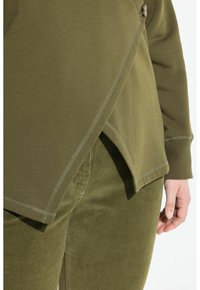 Primo piano di una mano in pantaloni in velluto a coste verde oliva e top a maniche lunghe con orlo asimmetrico abbinato, con cuciture visibili e dettaglio dei bottoni.