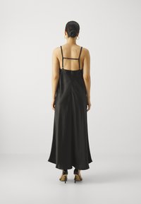 Svart sateng maxi-kjole med tynne stropper, åpen rygg og flytende kant. Brukt med svarte hælsko. Minimalistisk design på en ensfarget bakgrunn.