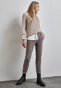 Beige fuzzy trui over een witte shirt, gecombineerd met smalle bruine broeken en zwarte enkellaarsjes op een grijze vloer.