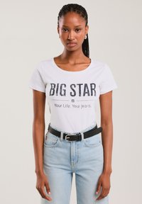 Žena s copánky, která má na sobě bílý nátělník s nápisem "BIG STAR Your Life. Your jeans." zastrčený do světle modrých džínů s černým páskem, neutrální pozadí.