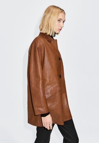 Brun læderjakke med løs pasform, stor krave, to knapper og en sidelomme. Teksturen er glat med en subtil glans.